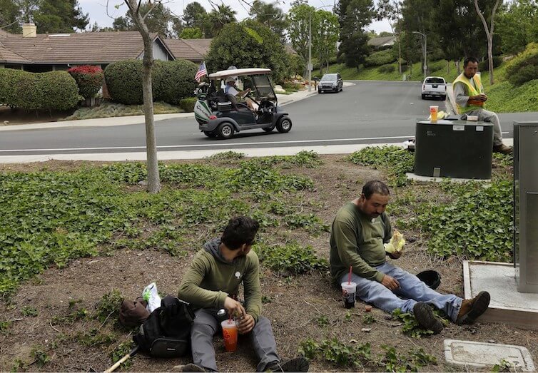 A man drives a golf cart from his house to his golf club as a group of landscape workers take a break in Vista, California.