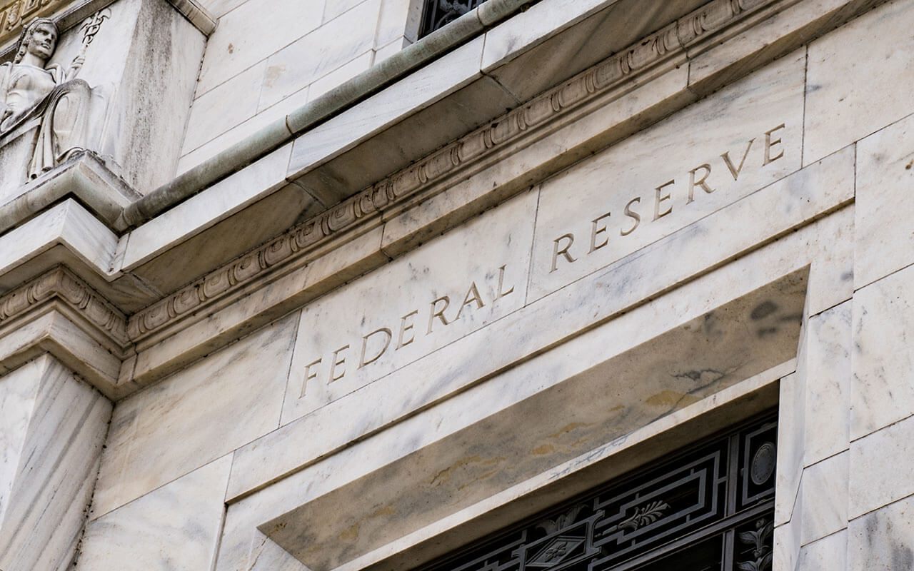  The image shows the exterior of the Federal Reserve building, which is made of marble and has the words "FEDERAL RESERVE" carved into the stone above the entrance.